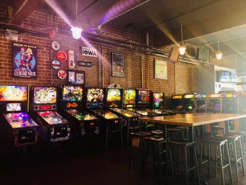 A cozy arcade with colorful pinball machines lined up against a brick wall and bar seating in a dimly lit space.