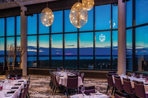 Elegant restaurant interior with round tables, large windows showcasing a sunset view over water, and decorative hanging lights.