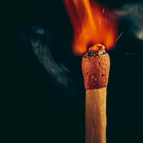 A close-up of a lit match with a bright flame and smoke against a dark background.