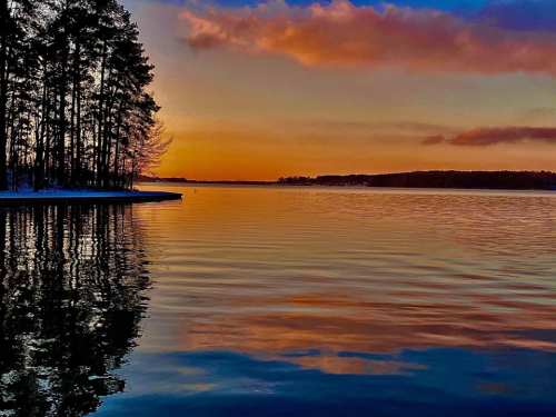 A serene lake at sunset, with colorful reflections and silhouetted trees along the shore.