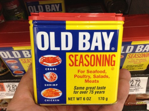 A hand holding a can of Old Bay seasoning, featuring crabs, shrimp, and chicken on a bright yellow and blue label.