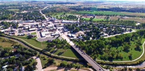 Aerial view of a small town with a river, green fields, and roads, surrounded by hills and open landscape.