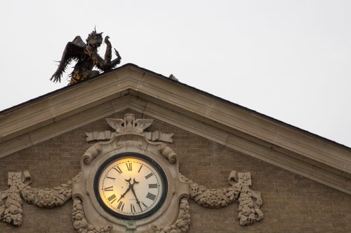 A clock on a building's facade with a decorative eagle statue perched on the roof above it.