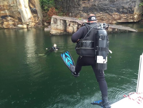 A diver prepares to jump into the water while another diver floats nearby in a rocky, green-tinted lake.