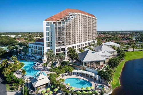 Aerial view of a large hotel with a pool area, surrounded by palm trees and a lake under a clear blue sky.