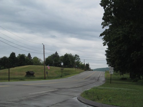 A rural road curves ahead, flanked by green hills and trees under a cloudy sky, with a small shed and an American flag visible.