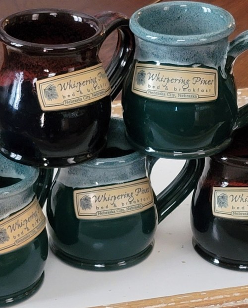 A stack of colorful ceramic mugs with "Whispering Pine Bed & Breakfast" labels, featuring dark red and green glazes.