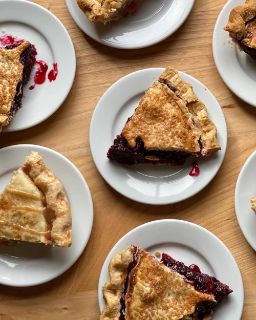 Slices of berry pie on white plates, arranged on a wooden table, with some pie filling oozing out.