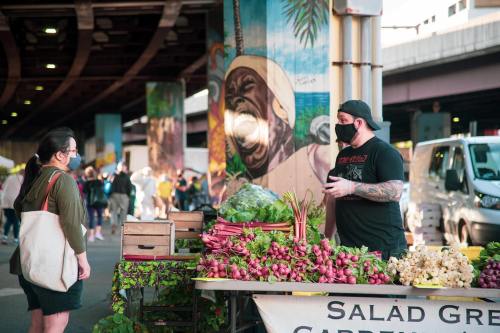 A farmer's market scene with a vendor selling fresh produce to a customer, colorful murals in the background.