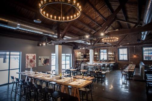 A spacious restaurant with high ceilings, wooden beams, and modern decor, featuring a long dining table and bar area.