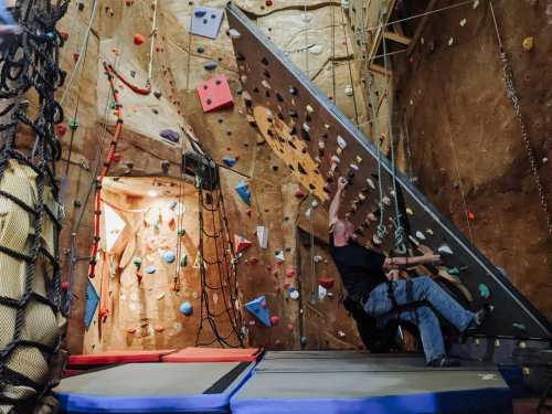 A person climbs a bouldering wall indoors, surrounded by colorful holds and safety mats.