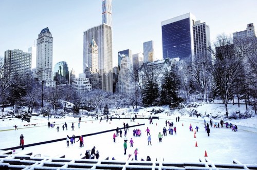 A bustling ice skating rink in a snowy park, surrounded by tall city buildings and trees.