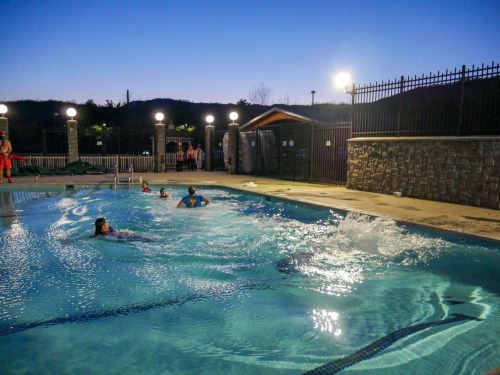 A pool at dusk with people swimming and splashing, surrounded by lights and a scenic backdrop of hills.
