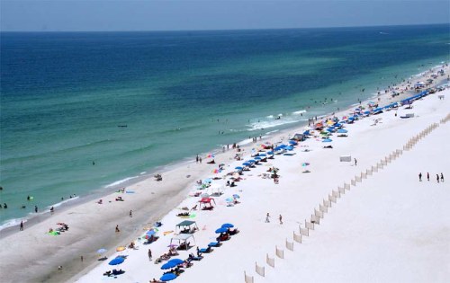 Aerial view of a sunny beach with colorful umbrellas, people swimming, and gentle waves along the shoreline.