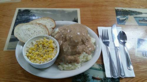 A plate of mashed potatoes topped with gravy, served with corn, two slices of bread, and cutlery on a wooden table.