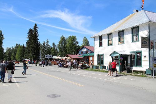 A bustling street scene with people walking, shops, and trees under a clear blue sky.