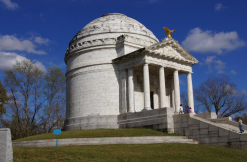 A white marble monument with a dome and golden eagle, surrounded by trees and a blue sky.