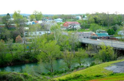 A scenic view of a small town with a river, greenery, and buildings, including a bridge and various shops.