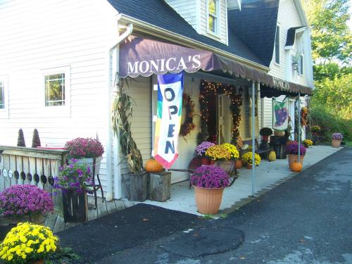 A charming shop entrance with a "Monica's" sign, colorful flower pots, and an "Open" banner.