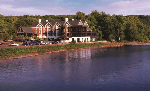 A scenic view of a riverside hotel surrounded by trees, with parked cars in front and a calm water surface.