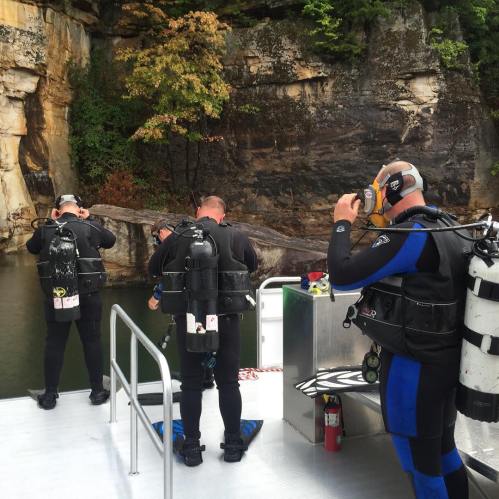 Divers in wetsuits prepare to enter the water from a boat near a rocky cliff and trees.