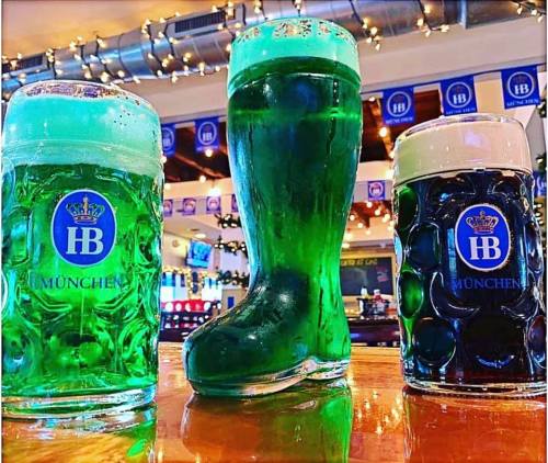 Three beer glasses on a bar: two traditional mugs and a large green boot-shaped glass filled with green beer.