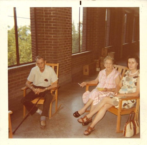 Three people sitting in rocking chairs in a brick-walled room, with large windows and natural light.