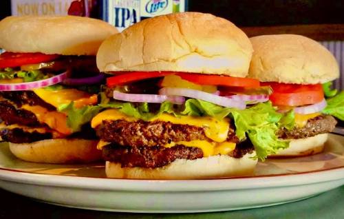 Three stacked cheeseburgers with lettuce, tomato, onion, and melted cheese on soft buns, served on a plate.