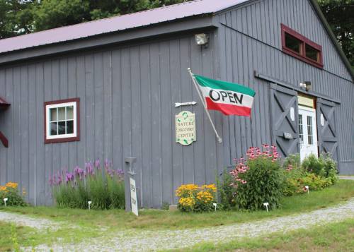 A gray barn with a green and red "OPEN" flag, surrounded by colorful flowers and a sign for a nature center.