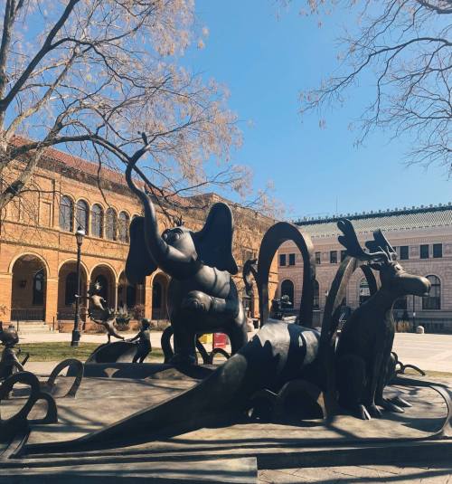 A large, abstract bronze sculpture featuring various animal forms, set against a clear blue sky and historic buildings.