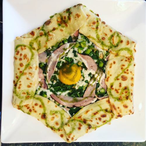 A hexagonal plate with flatbread surrounding a mix of greens, ham, cheese, and a raw egg yolk in the center.