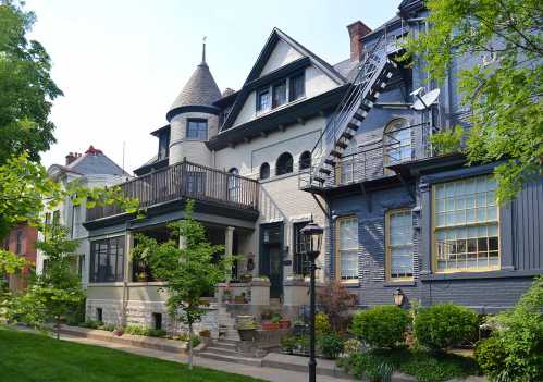 A charming row of historic houses with varied architectural styles, featuring a balcony and a fire escape. Lush greenery surrounds.