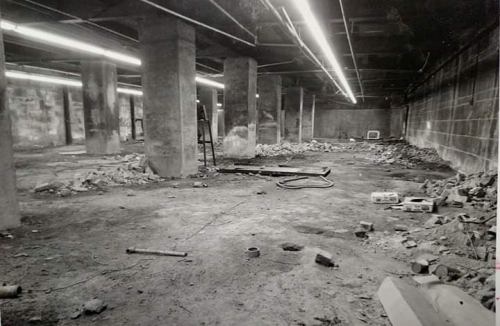 A dimly lit, empty basement with concrete pillars, debris, and scattered construction materials on the floor.