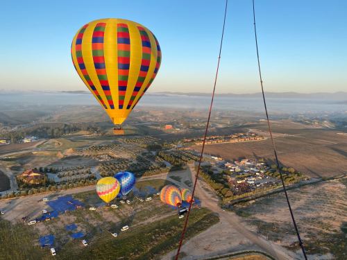 A colorful hot air balloon floats above a landscape with other balloons and a parking area below.