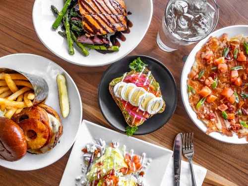 A table spread with grilled chicken, fries, a burger, nachos, and a salad, accompanied by a glass of water.