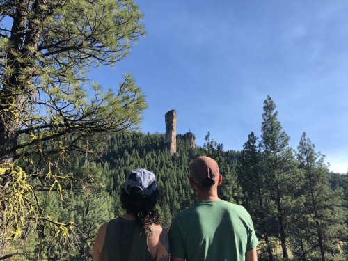 Two people stand facing a tall rock formation surrounded by lush green trees under a clear blue sky.