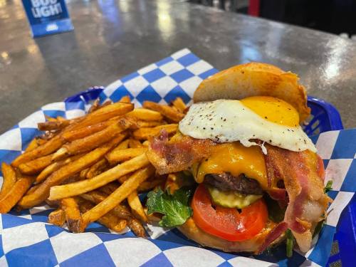 A juicy burger topped with bacon, cheese, and a fried egg, served with a side of golden French fries in a blue checkered basket.