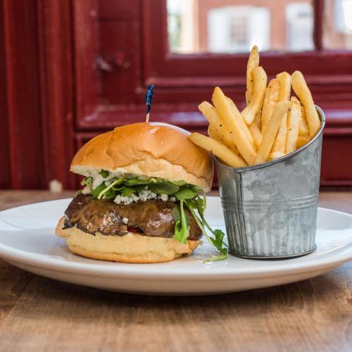 A gourmet burger with greens and blue cheese, served with a side of crispy French fries in a metal cup.