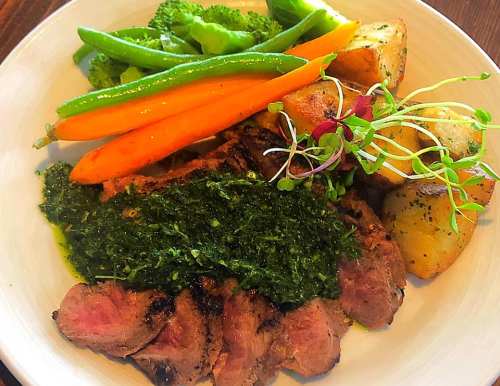 A plate of grilled steak with green sauce, served with broccoli, green beans, carrots, and roasted potatoes.