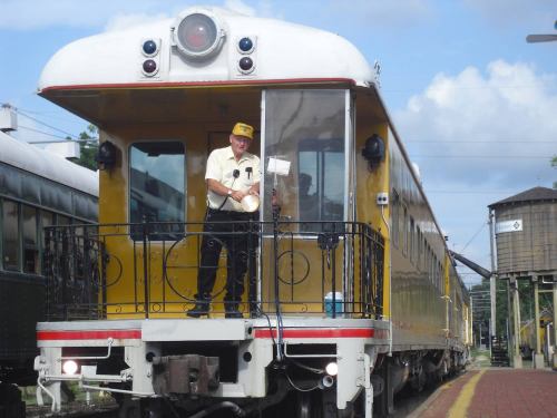 A smiling conductor stands at the front of a vintage yellow train, ready to depart on a sunny day.
