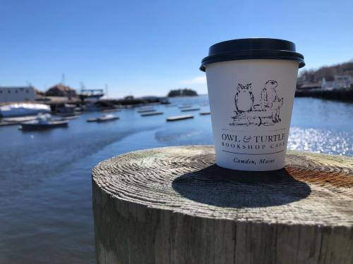 A coffee cup on a wooden post by a harbor, with boats and a clear blue sky in the background.