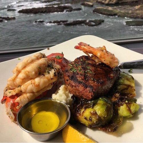 A gourmet plate featuring grilled shrimp, steak, Brussels sprouts, and a lemon wedge, with a scenic ocean view.