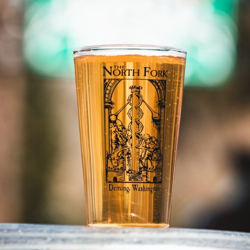 A clear glass of beer with "The North Fork" logo, set against a blurred background.