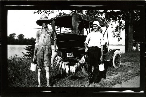Two men stand beside an old car, holding buckets and displaying fish, with a river and trees in the background.