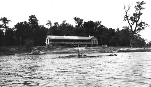 A black and white photo of a river with a rowing boat in the foreground and a building surrounded by trees in the background.