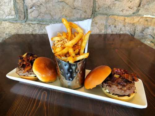 Two mini burgers with bacon and a side of crispy fries in a metal cup, served on a white plate against a stone wall.