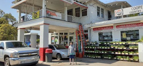 A man stands outside a two-story store with a sign for electrical supplies and a variety of plants for sale.