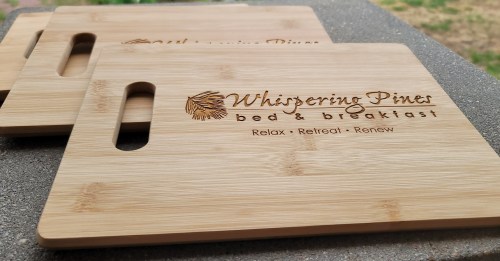 Three wooden cutting boards with "Whispering Pines bed & breakfast" engraved, featuring the slogan "Relax, Retreat, Renew."