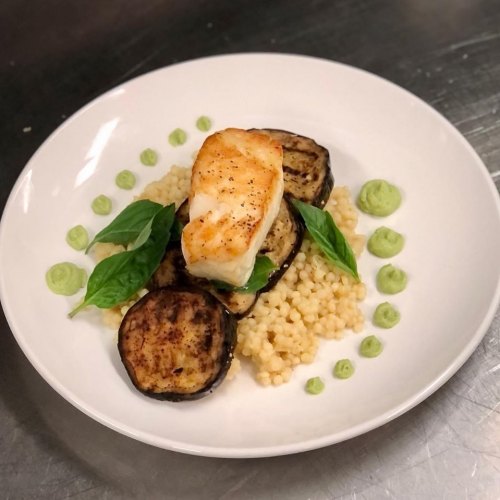 A plate of grilled fish atop couscous and eggplant, garnished with basil and green sauce dots.