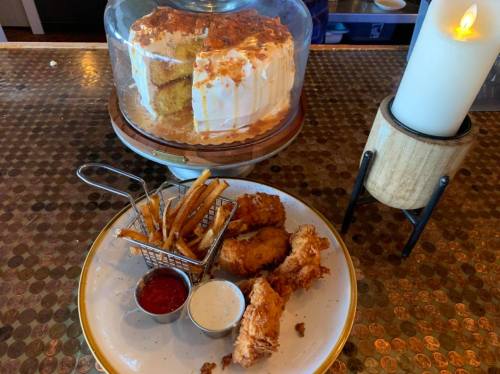 A plate with fried chicken, fries in a small basket, and dipping sauces, with a cake displayed in the background.